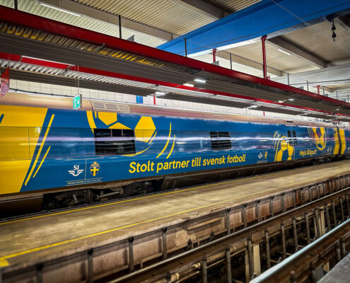 Tågfoliering av SJ X55 för fotbolls-VM 2026 Helfolierad SJ-vagn i blått och gult med fotbollsmotiv och texten Stolt partner till svensk fotboll